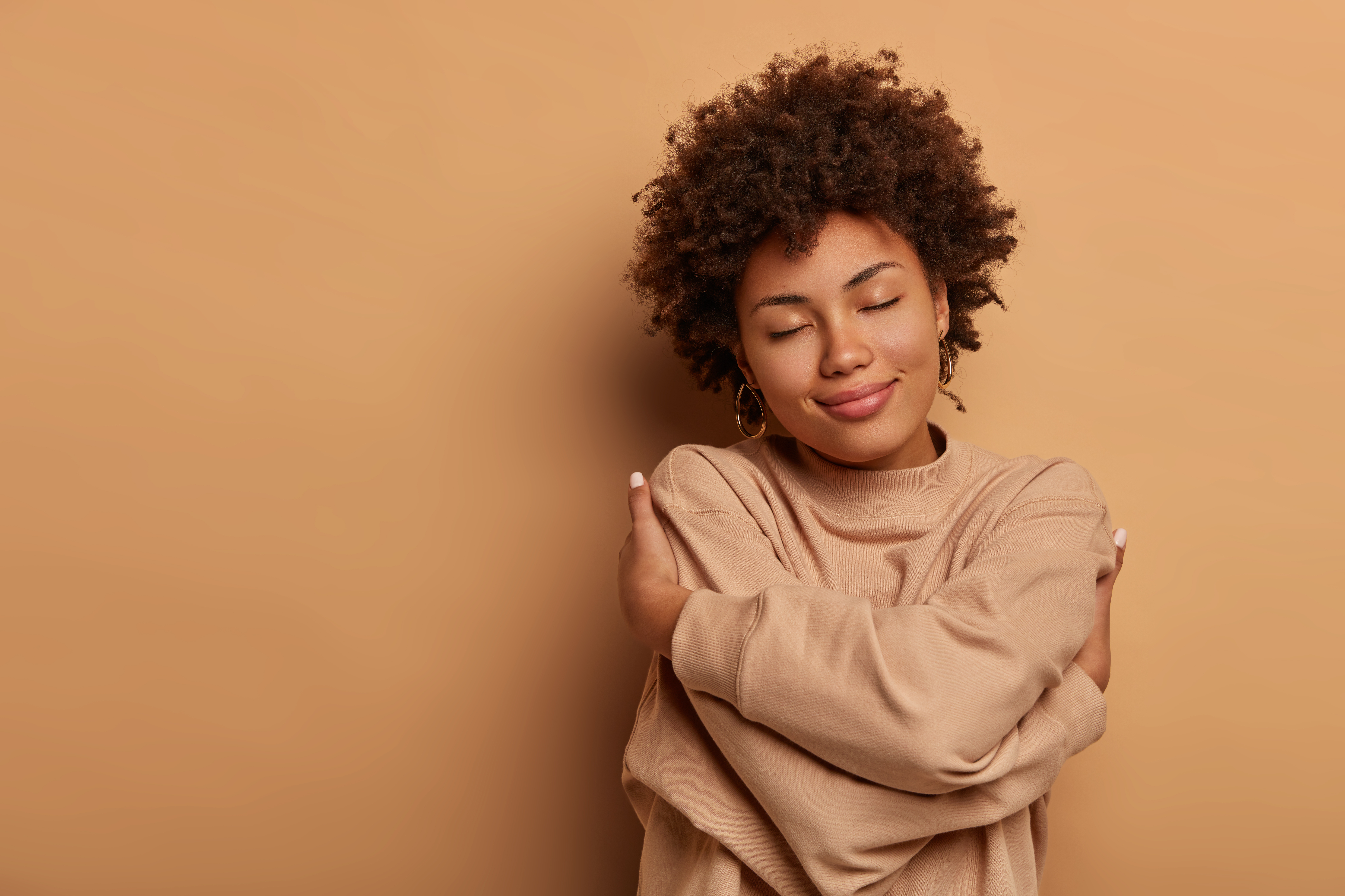Young black woman with short, curly hair in tan sweater gives herself a hug with eyes closed against tan gradient background.