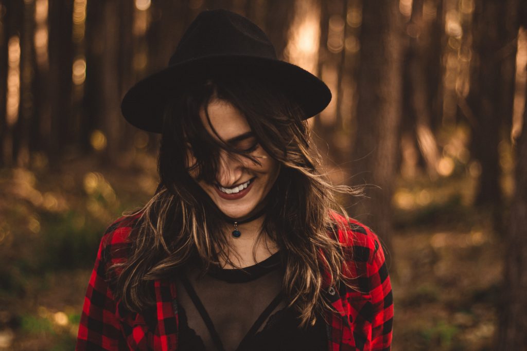 Young white woman with long brown hair in black fedora and red and black checkered shirt, looking downward and smiling as she walks through the woods.