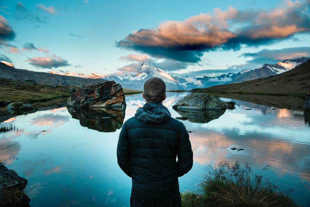 A man with short hair and a dark winter jacket facing a spacious landscape of lake, mountains and sky.