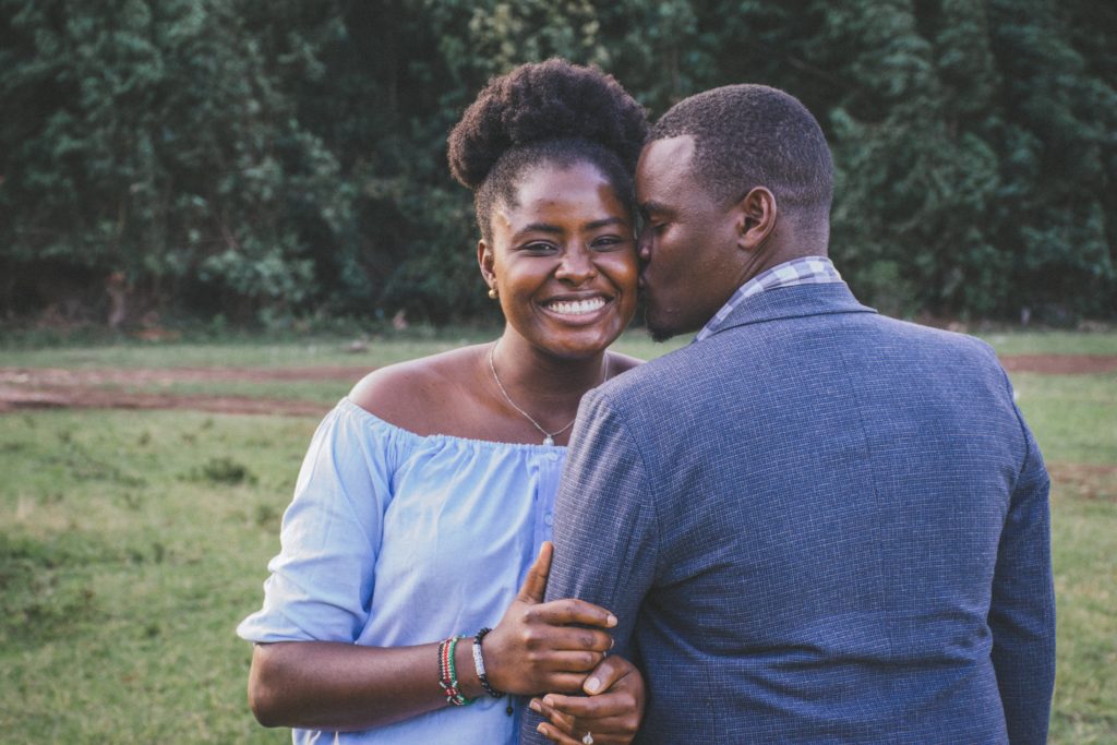 Young smiling Black woman wearing an off-the-shoulder blue top being kissed on the left cheek by a young Black man in a blue sport jacket, against a background of green trees.