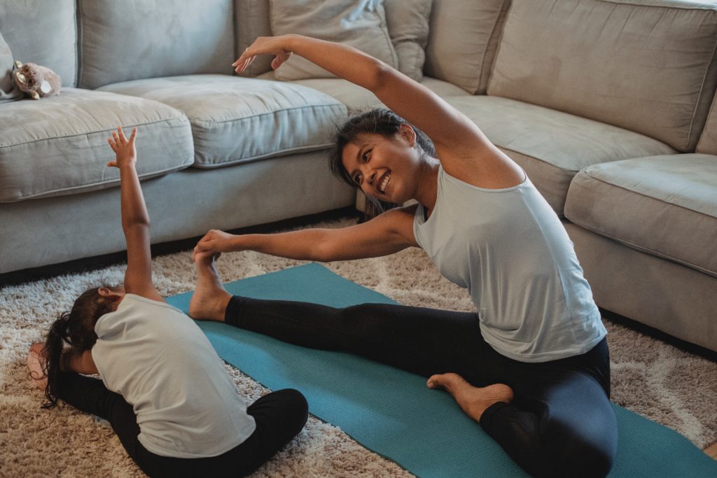 Young Asian mom in a sleeveless grey top and leggings side-stretching and smiling on a yoga mat, facing her young daughter who is doing the same.
