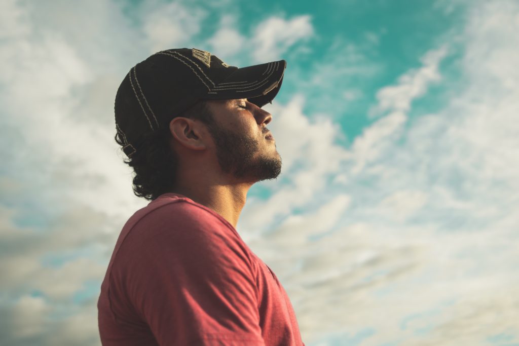 Man with short beard and black ball cap with eyes closed and head tilted toward the blue sky.