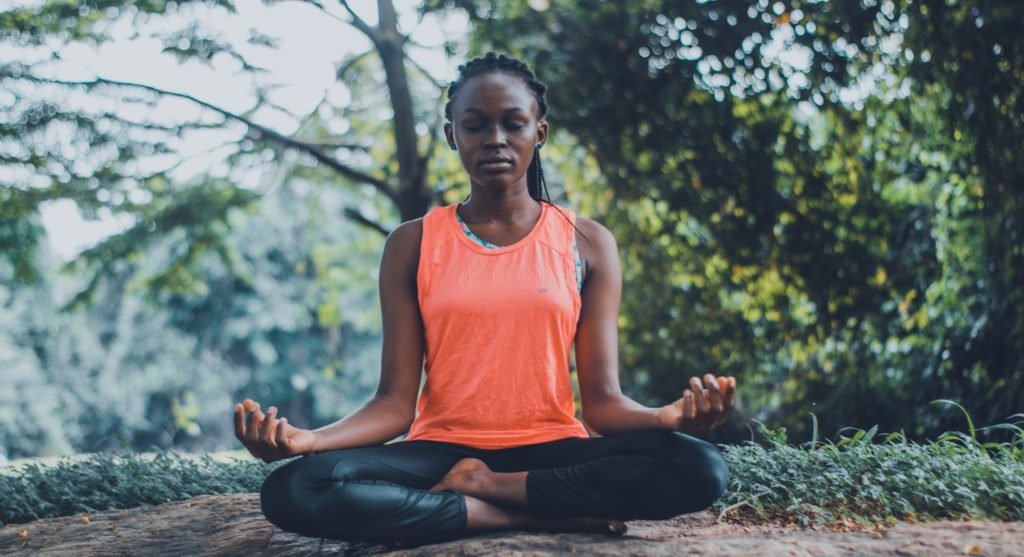Woman meditating outdoors, wearing an orange sleeveless shirt and leggings, seated in a cross-legged position.